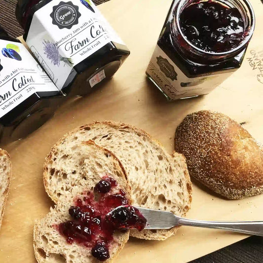 Our Blueberry with a Hint of Lavender Jam – One Open Jar and Spread on a Plate of Sourdough.