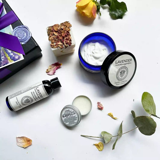 Close-up view of the Beautiful Hand & Massage Therapy Gift Box in a black gift box with a purple ribbon, showcasing an open lid of lavender hand cream and sleep balm to reveal textures, along with lavender massage oil and handcrafted lavender essential oil soap, decorated with fresh yellow roses, eucalyptus leaves, and rose petals.