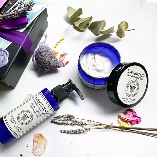 Close-up view of the Lavender Beauty Skincare Gift Set in a black gift box with a purple ribbon, featuring an open lid of lavender hand cream to reveal its texture, alongside lavender silk lotion and a dried lavender sachet. Background decor includes dried lavender flowers, rose petals, and eucalyptus leaves.