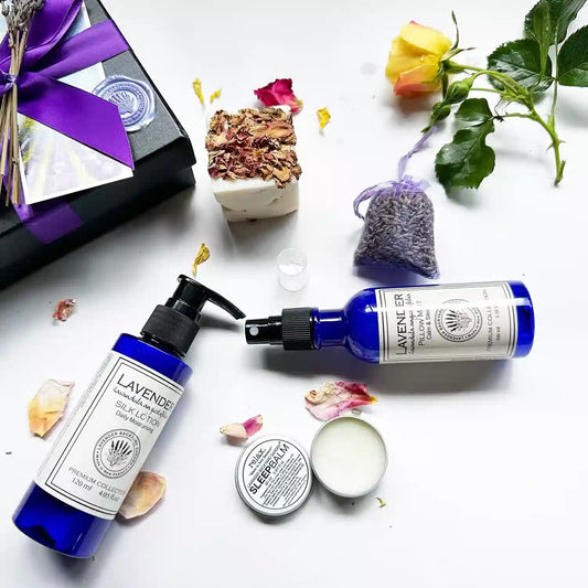 Close-up view of Lavender Wishes Holiday Gift Ideas, featuring a Small Lavender Sachet Bag, Lavender Pillow Mist, Lavender Silk Lotion, open Lavender Sleep Balm showing texture, and Lavender Essential Oil Handmade Soap (assorted), alongside a black gift box with a purple ribbon, decorated with yellow roses and rose petals.