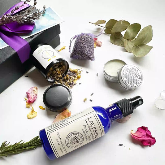 Close-up view of Lovely Lavender Therapy Giftset for Women featuring dried lavender sachet, lavender sleep balm (open lid to show balm texture), lavender pillow mist, and lavender & chamomile herbal tea - loose leaf (travel size, with open lid showing chamomile and dried lavender herbs), set against a background of eucalyptus leaves, rosemary, and rose petals.