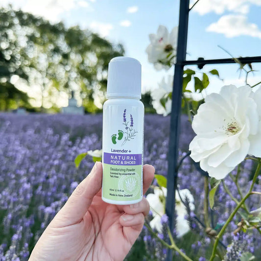 Natural Foot & Shoe Powder for Smelly Feet, held by hand against a purple lavender field beside white roses.