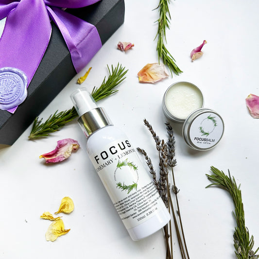 Close-up of "Lavender & Rosemary Focus Balm, Focus Aromatherapy Spray with Pure Essential Oils" with rosemary leaves, dried lavender flowers, and rose petals as background decor with a purple ribbon gift box.