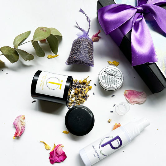 Close-up view of the "Thoughtful Small Gifts for Teachers," featuring Lavender Sleep Balm Essential Oil Blends, Lavender Sleep Spray (Travel Size), Lavender & Camomile Herbal Tea (Mini) with the open lid revealing chamomile and lavender dried flowers, and a Dried Lavender Sachet. The background includes rosemary, rose petals, and a black gift box with a purple ribbon.