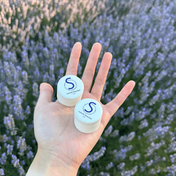 Two pots of Lavender Sleep Balm held by hand against a backdrop of blooming purple lavender