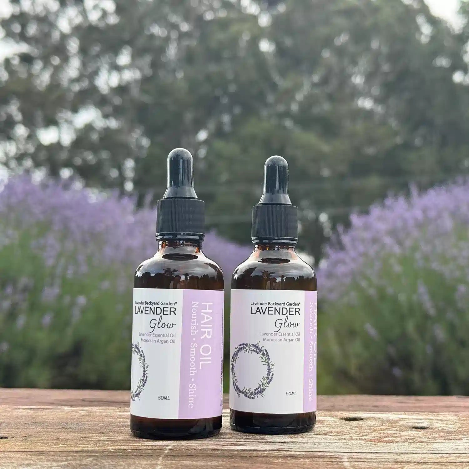 Two bottles of lavender hair oil placed on a wooden bench against blooming lavender plants.