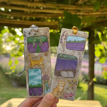 Hands holding two lavender scented walking cat bookmarks in front of kiwifruit vines with a lavender field in the background